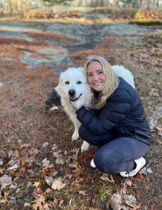 A young woman smiling and hugging a large white dog while crouching on a grassy area with fallen leaves.